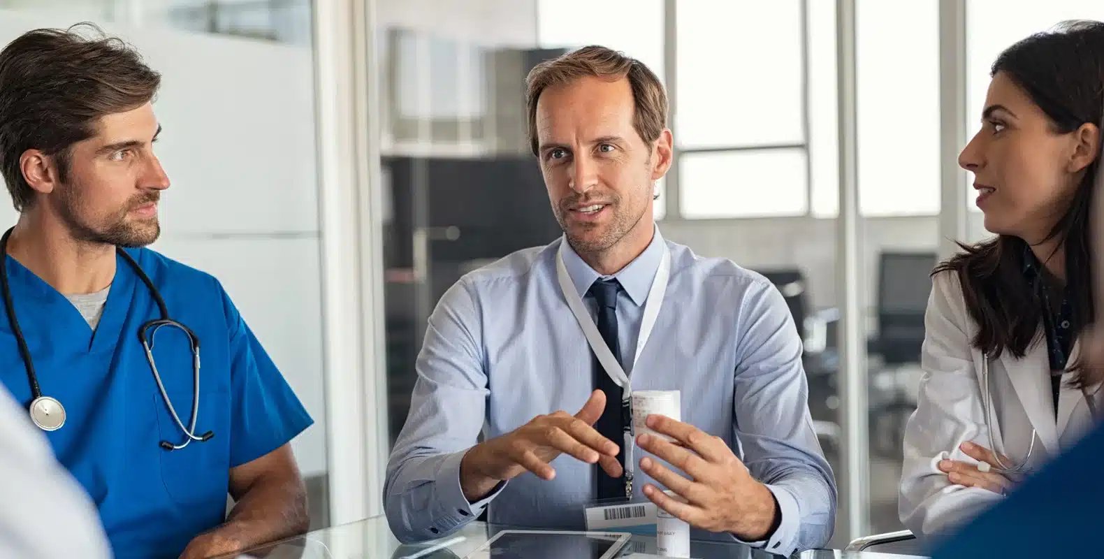 Three medical professionals in discussion at a table, including a doctor in blue scrubs and two in white coats.