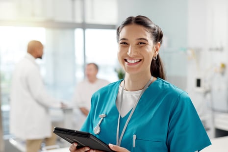 A smiling nurse in teal scrubs holding a tablet stands in a bright hospital room, representing the vital role of Allied Health professionals, while two healthcare experts discuss Medical Support Degrees in the background.