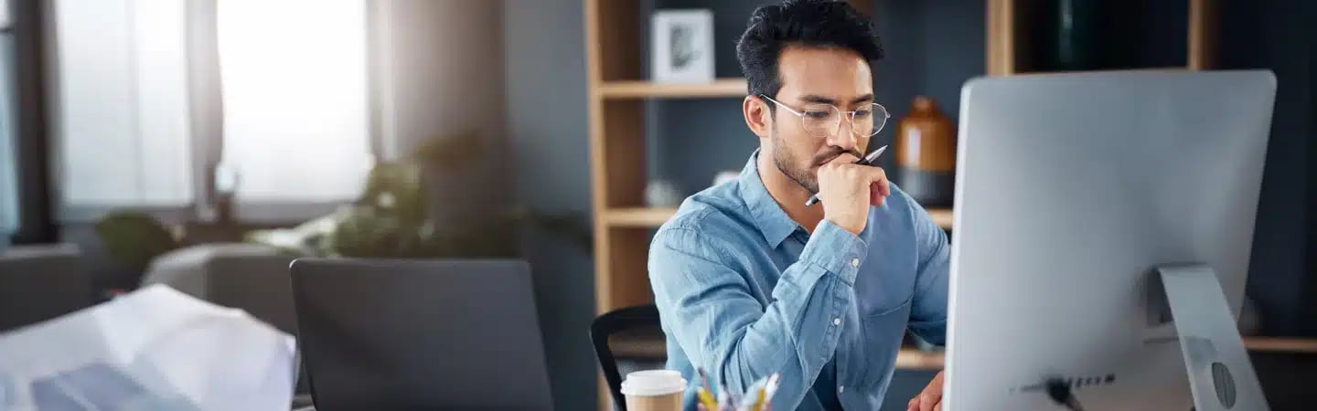 Man with glasses working at a computer in a modern office setting with shelves and papers in the background.