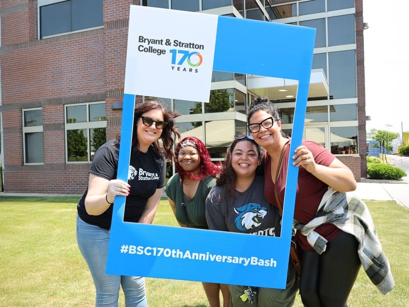 Four people smile while posing with a large blue frame outdoors. The frame reads "Bryant & Stratton College 170 Years" and "#BSC170thAnniversaryBash" at the bottom. A brick building with large windows is in the background.