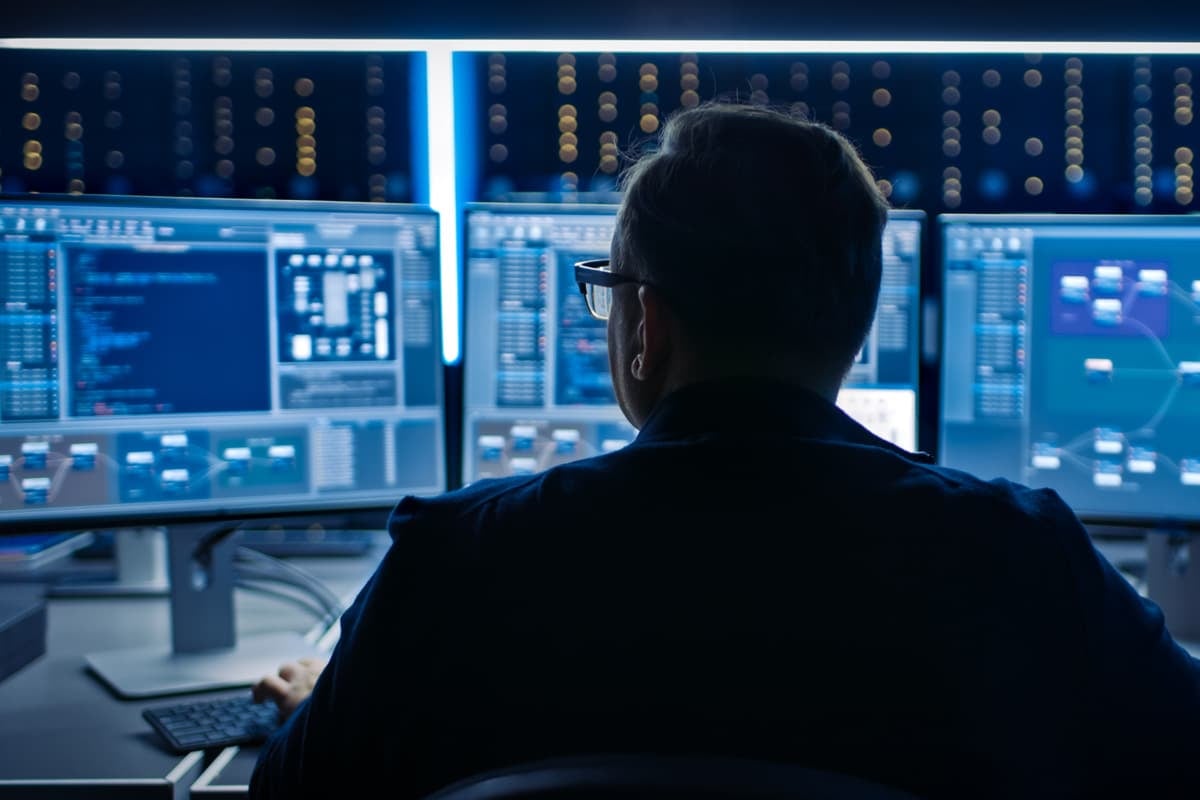 A person sits in front of multiple computer screens displaying various data and charts in a dimly lit room.