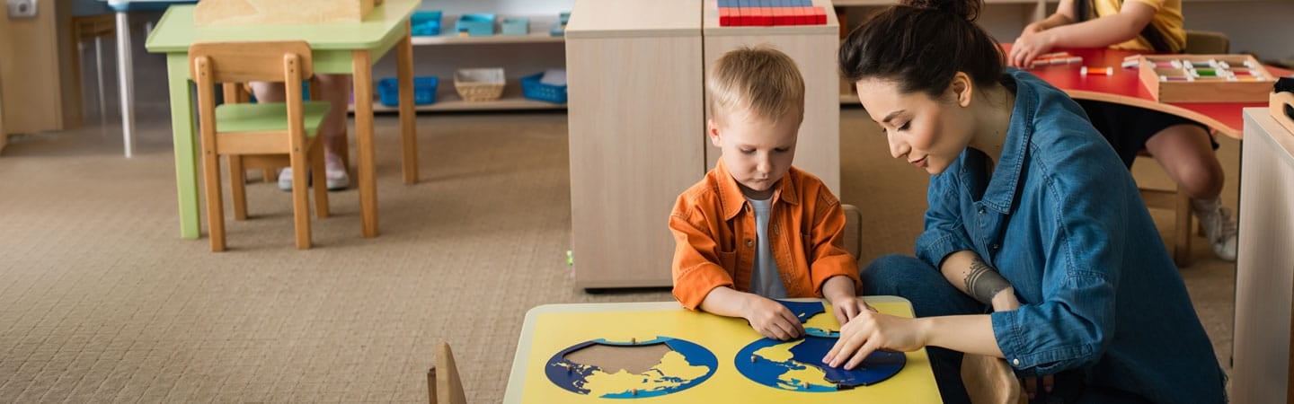 A woman assists a young child with a globe puzzle in a classroom setting. Other children are engaged in activities nearby.