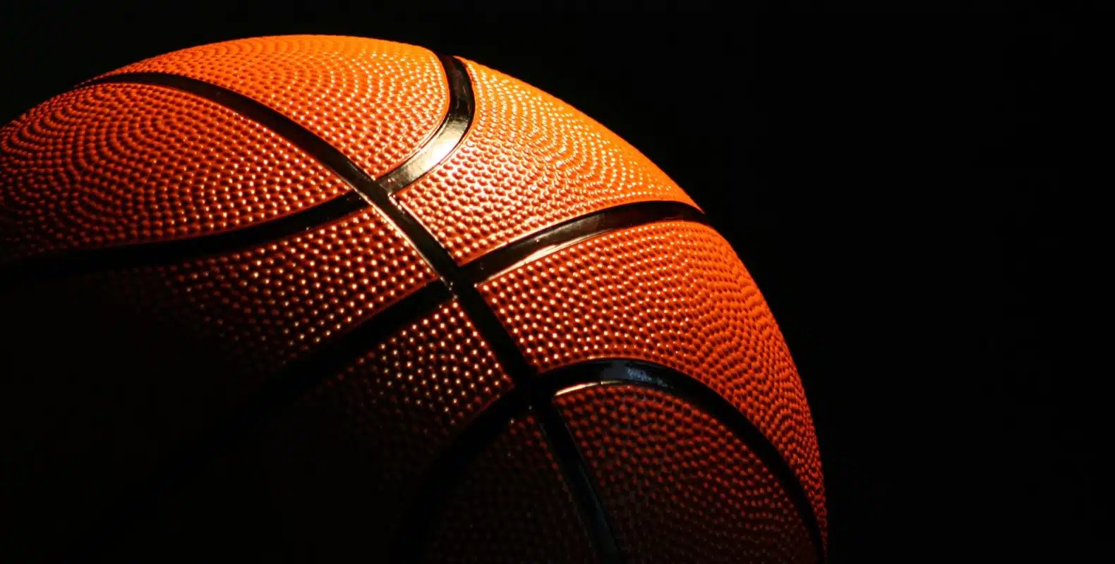 Close-up of a basketball with dim lighting, highlighting its textured surface and lines against a black background.