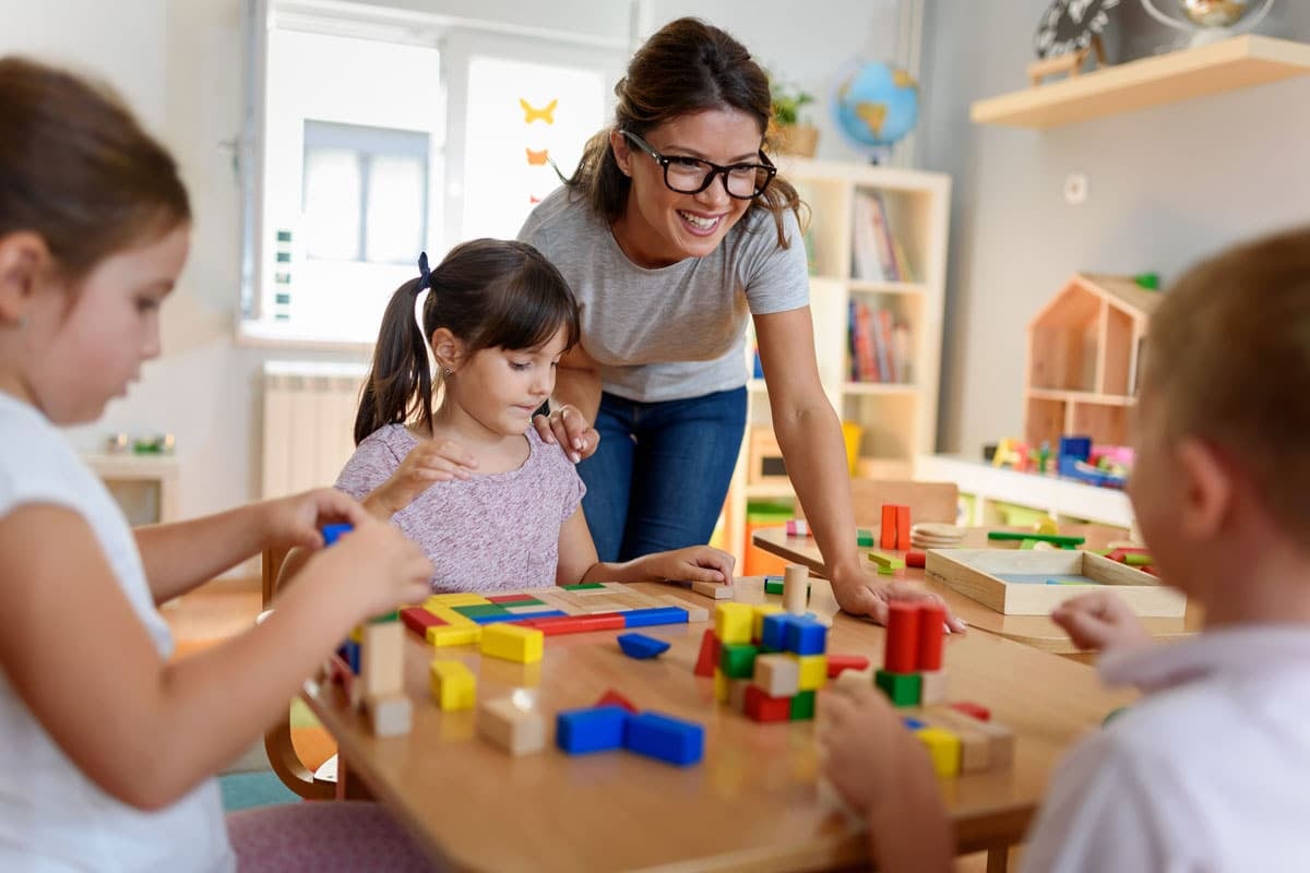 A teacher helps children playing with colorful blocks at a table in a bright classroom.