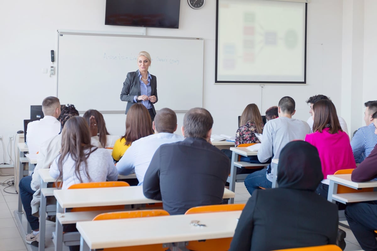A person stands at the front of a classroom, teaching a group of students sitting at white desks.