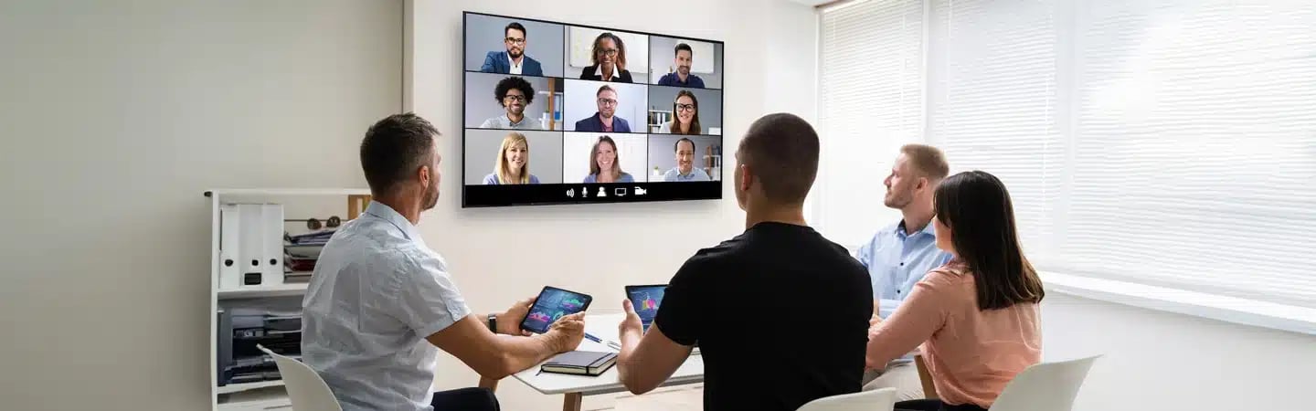 Four people in an office watch a video conference on a large screen, each using tablets and laptops.