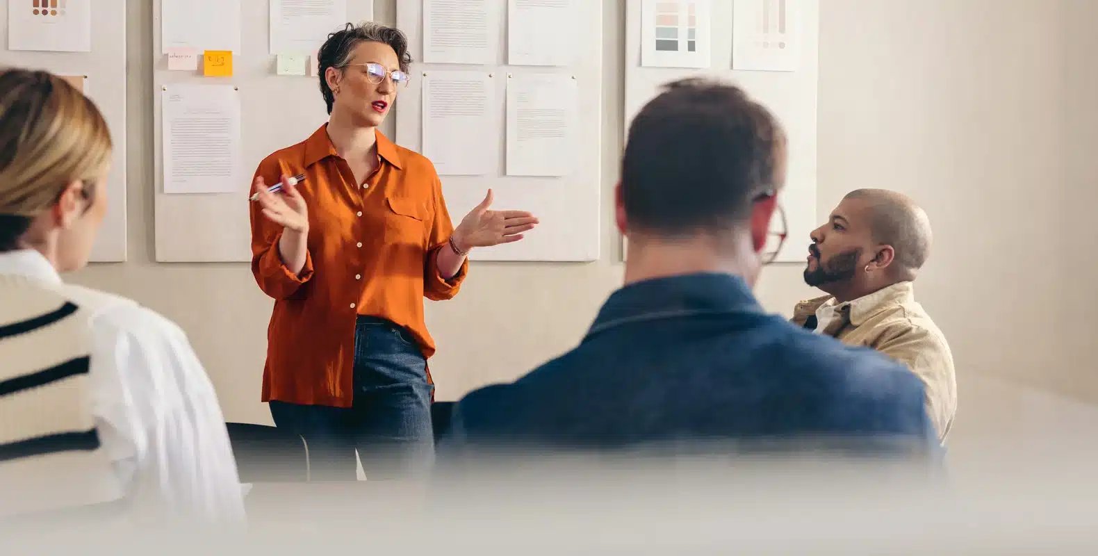 Person in orange shirt giving a presentation to a seated group in a meeting room with papers on the wall behind them.