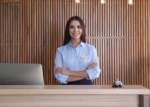 A woman in a blue uniform stands behind a reception desk with arms crossed, smiling. There is a laptop and a bell on the desk, and the background features vertical wooden paneling.