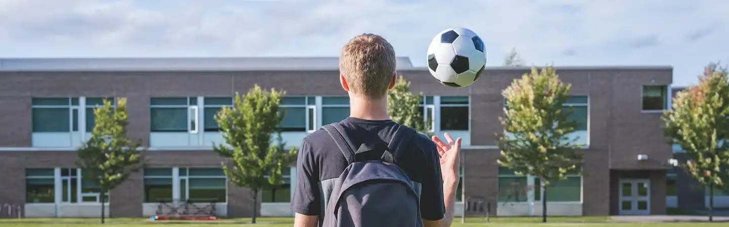A person with a backpack tosses a soccer ball in the air, standing in front of a school building and trees.