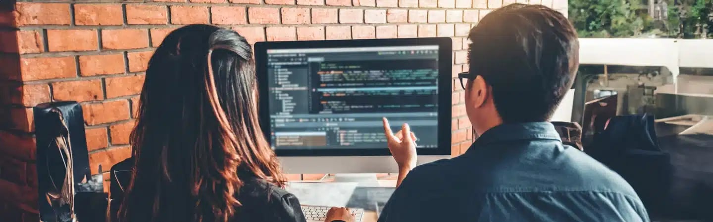 Two people working on a computer with code on the screen, sitting at a desk with a brick wall in the background.
