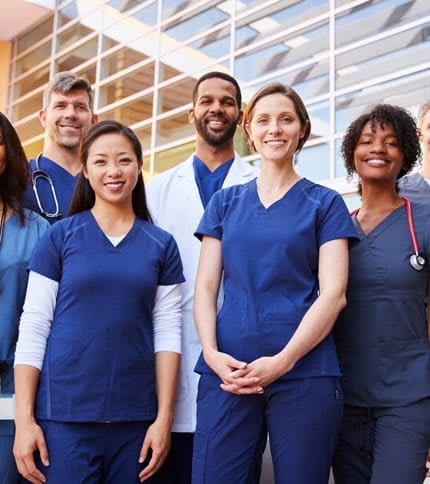 A group of healthcare professionals, wearing blue and white medical scrubs, stand smiling in front of a modern building with large windows.