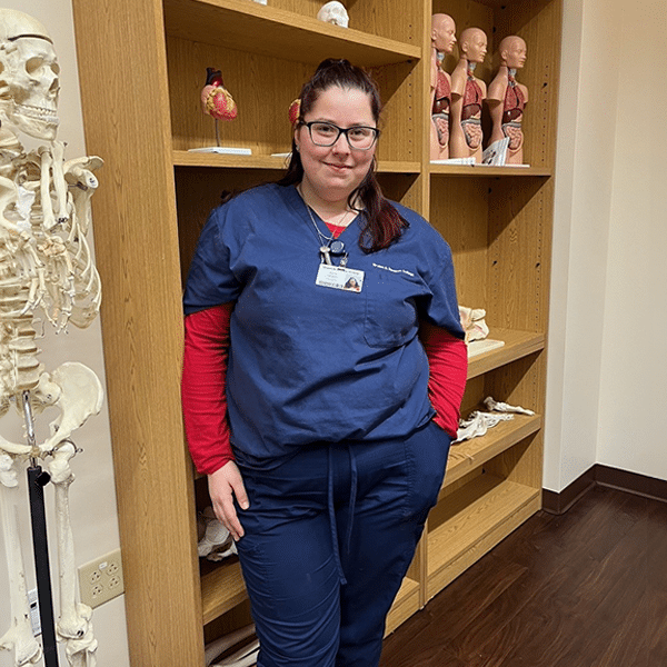 A person in navy scrubs and glasses stands in front of anatomical models and a skeleton in a medical classroom.