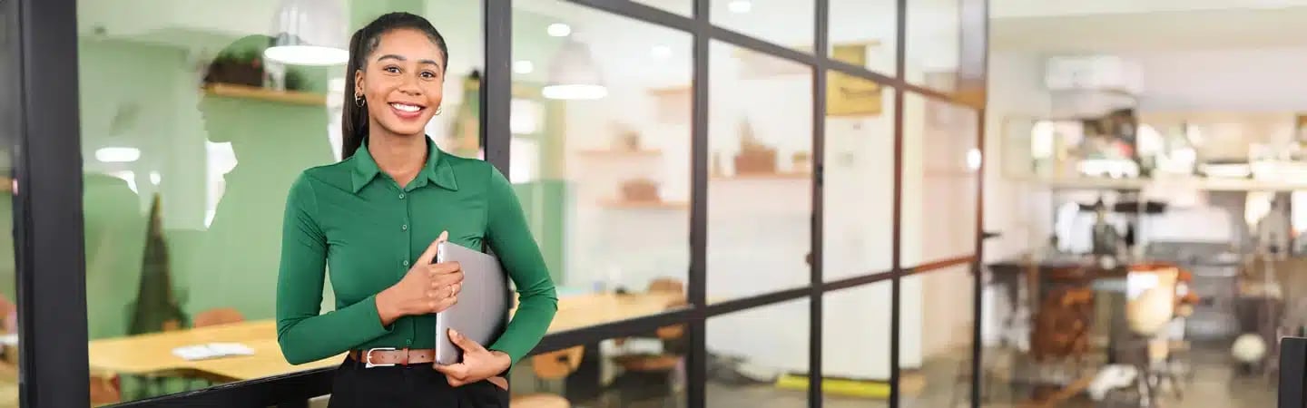 Smiling woman in a green shirt holding a tablet, standing in front of a glass-walled office.