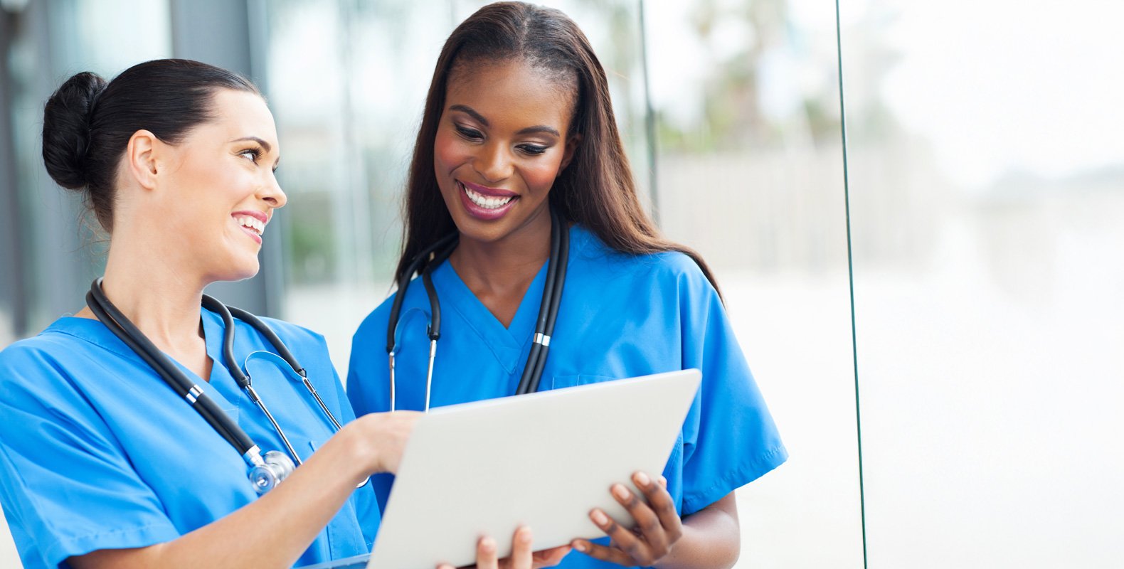 Two smiling healthcare professionals in blue scrubs and stethoscopes review a digital tablet together, possibly preparing for an upcoming CCNE accreditation review, while standing by a large window in a bright, modern setting.