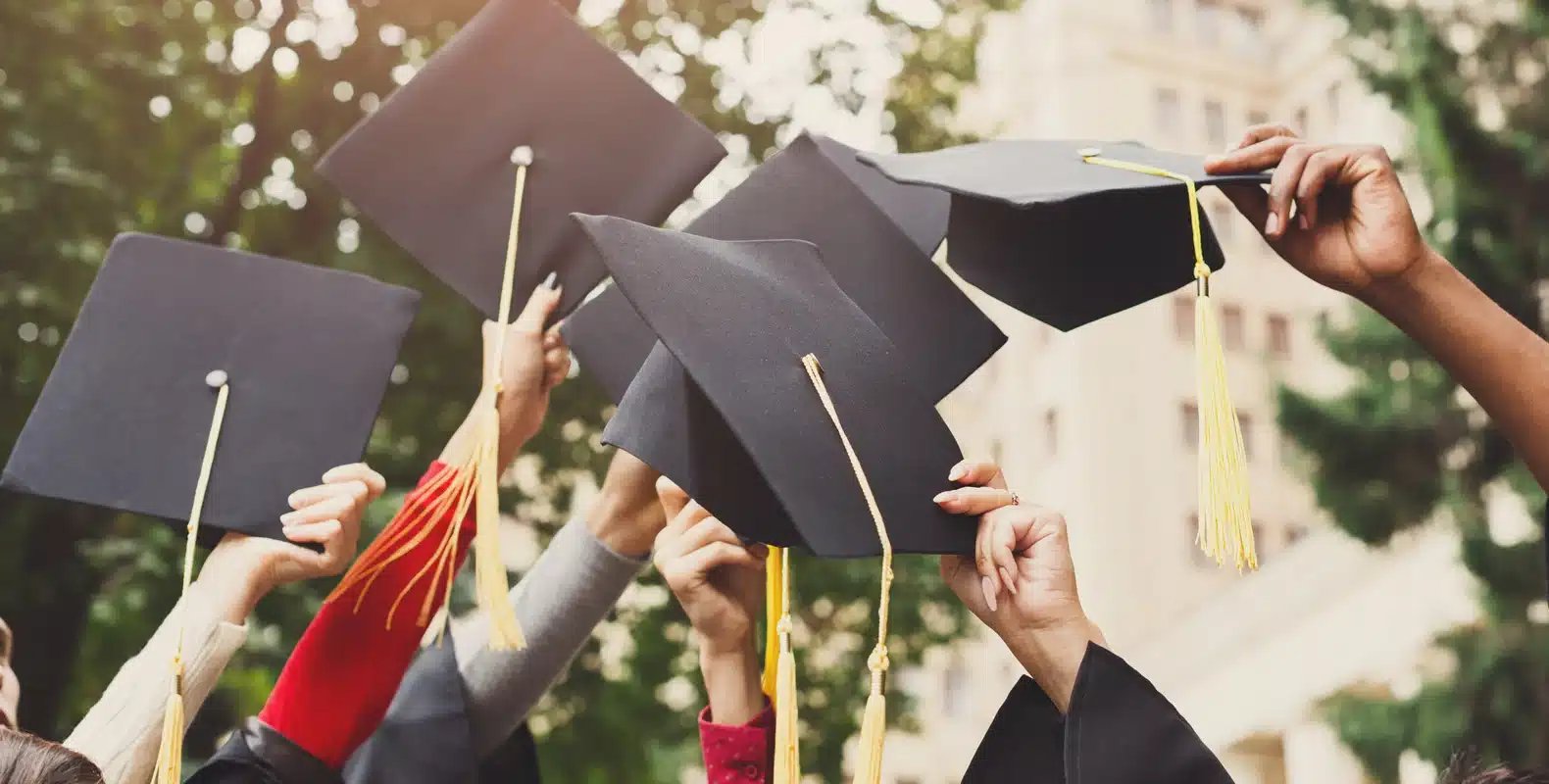 Graduates raise their mortarboards in the air, celebrating their achievement outdoors with trees and a building in the background.