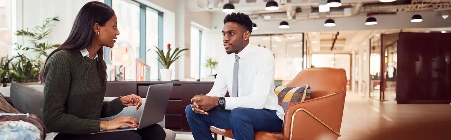 Two colleagues discuss work while seated in a modern office; one uses a laptop, the other listens attentively.