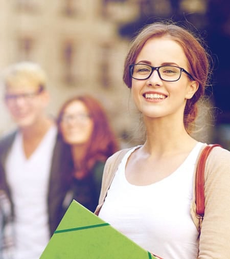 A young woman with glasses smiling and holding folders stands outdoors, with two people blurred in the background.