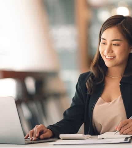 A woman in a blazer sits at a desk, smiling while working on a laptop. Open notebooks are in front of her, and the background is softly blurred, suggesting a modern office environment.