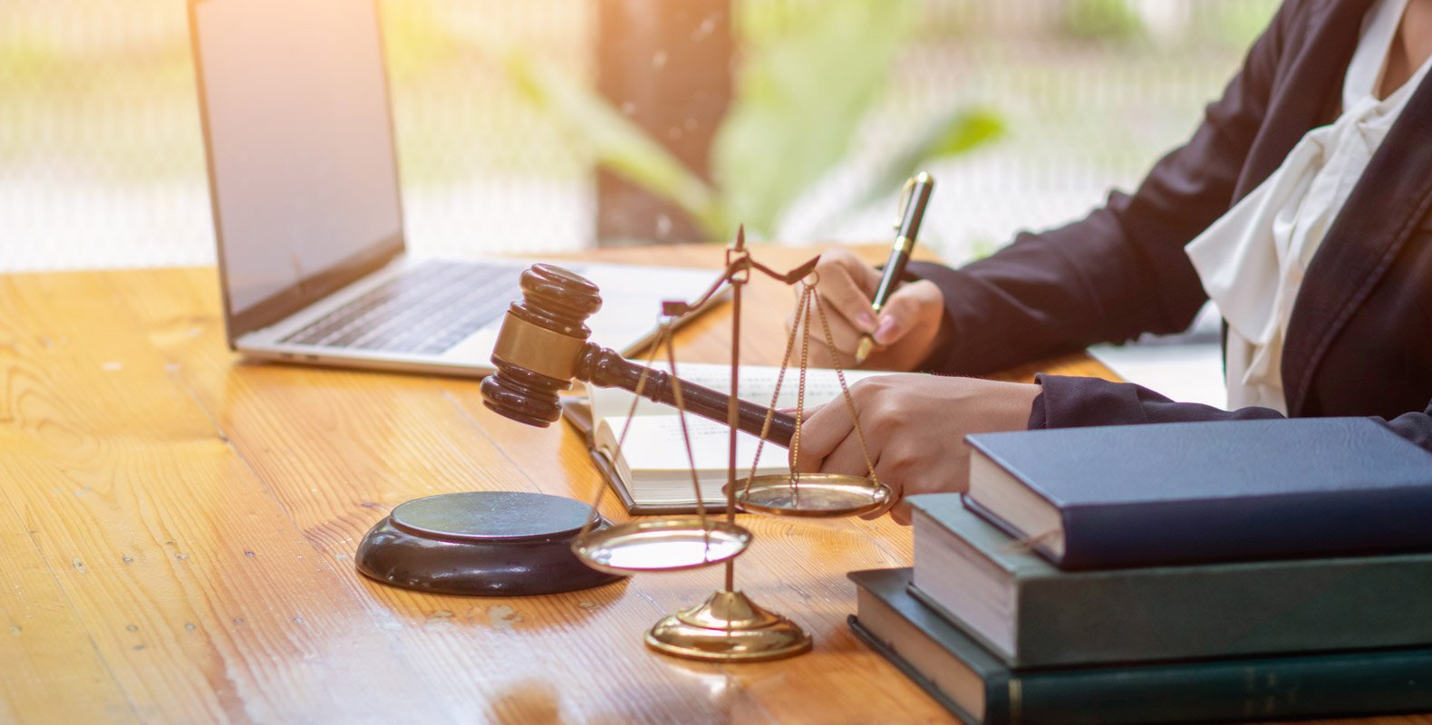 A person in professional attire sits at a desk with a laptop, writing in a notebook. A gavel, scales of justice, and stacked books are also on the desk, highlighting the work of a paralegal and symbolizing law and legal duties.