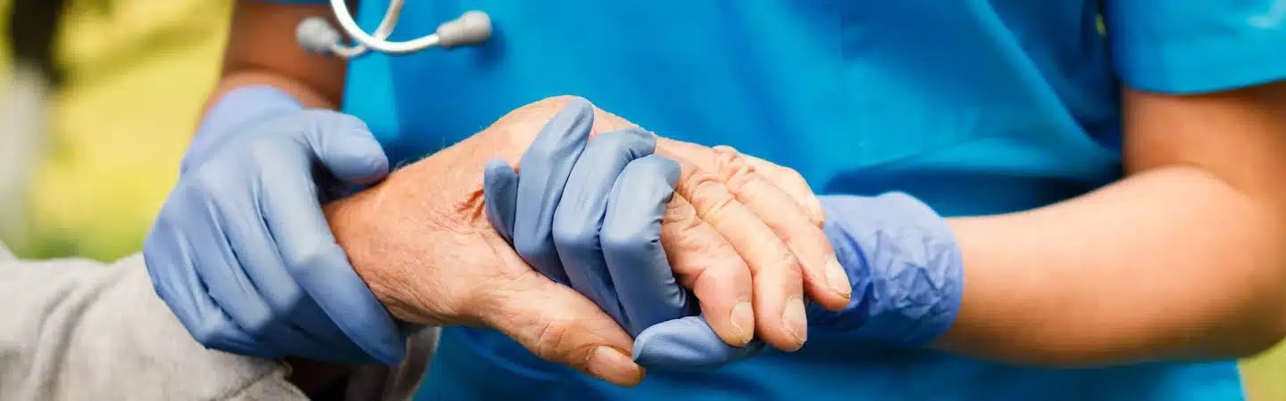 A healthcare worker in blue scrubs holds the hand of an elderly person, both wearing blue gloves.