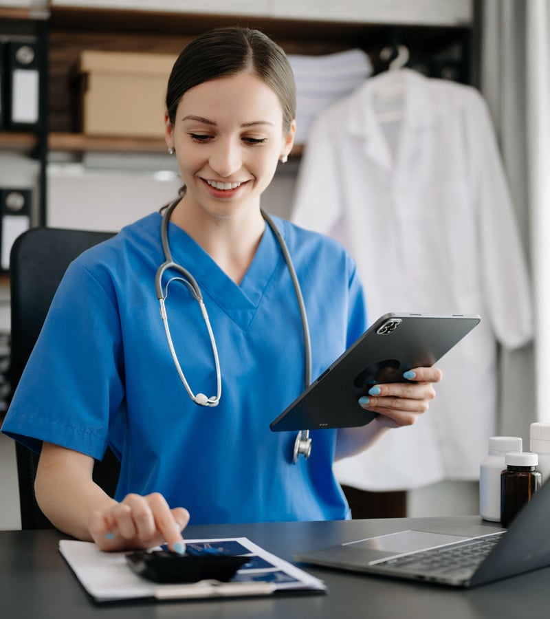 A smiling healthcare professional in blue scrubs with a stethoscope holds a tablet and reviews paperwork at a desk with medicine bottles, reflecting the vital role of medical coding in a busy healthcare office setting.
