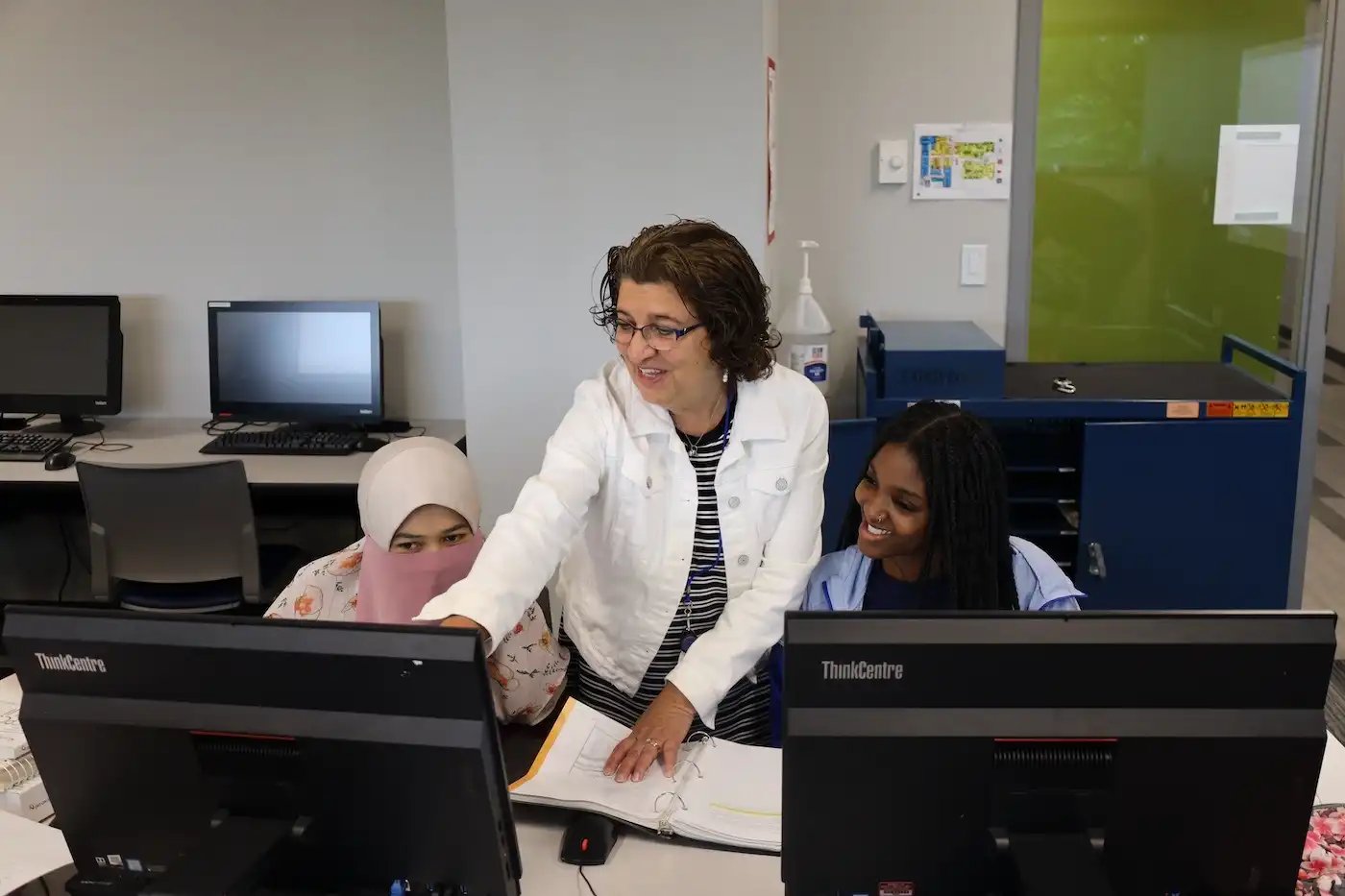 A woman points at a computer screen, helping two people who are sitting at desks in a classroom setting.