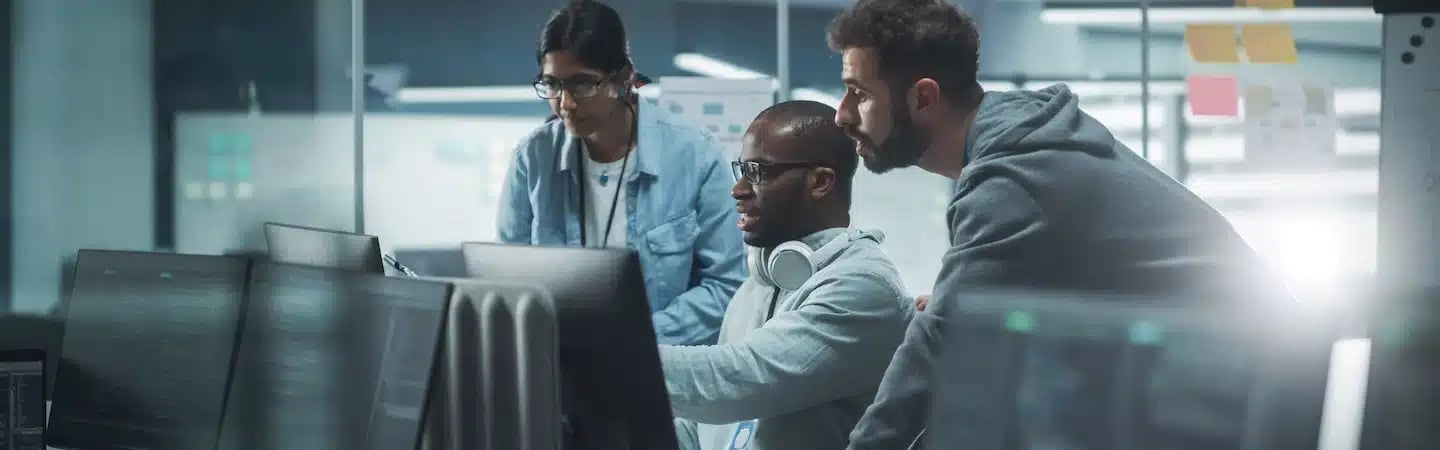Three people at a desk, looking at computer monitors in a modern office with glass walls and sticky notes.