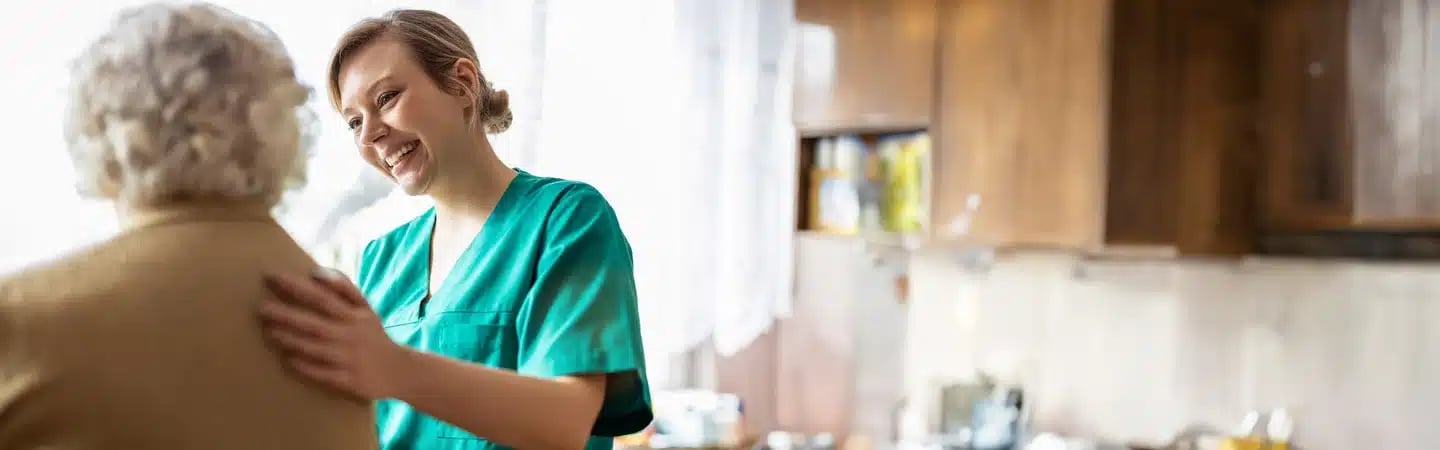 A caregiver in green scrubs smiles and comforts an elderly person in a home setting.