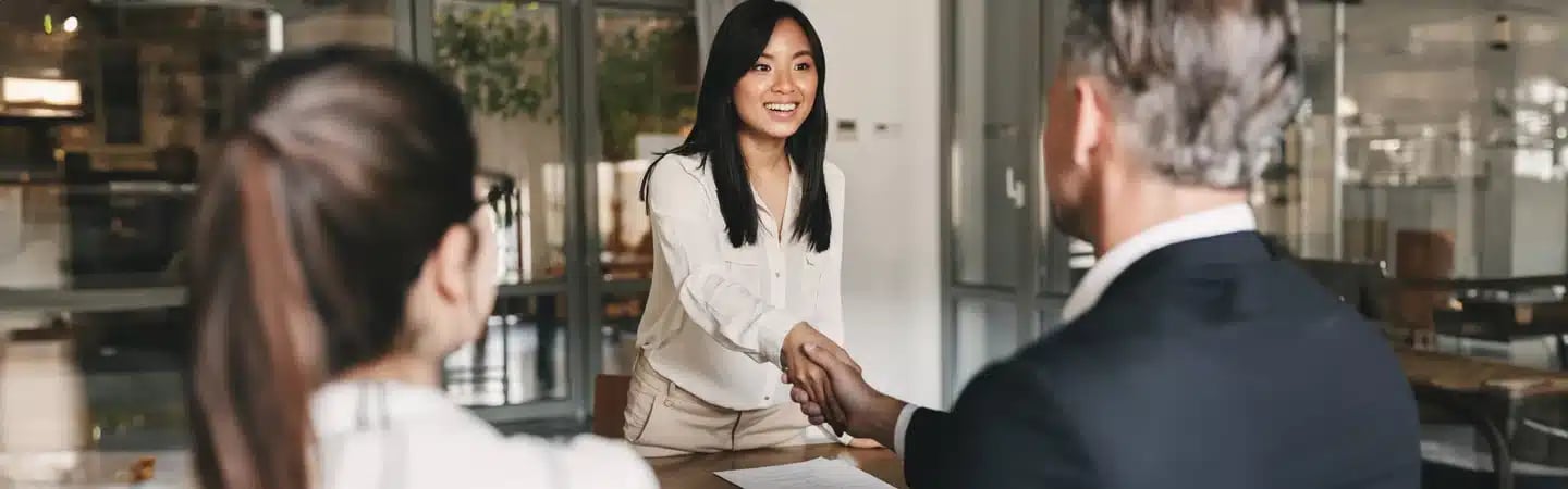 Woman smiling and shaking hands with a man across a table in an office setting. Another woman sits nearby, back to camera.