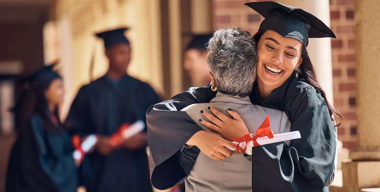Graduates in caps and gowns celebrate; one hugs an elderly person while holding a diploma. Other graduates chat in the background.