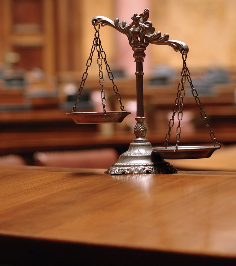 A close-up of a decorative brass balance scale on a polished wooden desk in a courtroom, with blurred chairs and walls in the background.