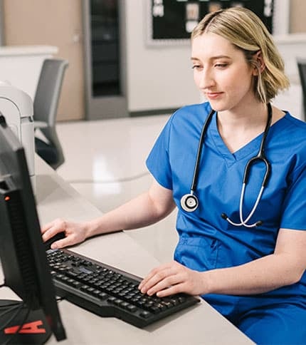 A healthcare professional in blue scrubs, with a stethoscope around their neck, uses their medical assistant associate degree as they work at a computer in a clinical setting.