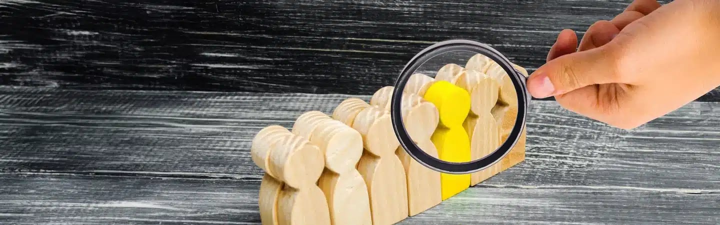 A hand holding a magnifying glass over wooden figures in a row, highlighting one yellow figure.