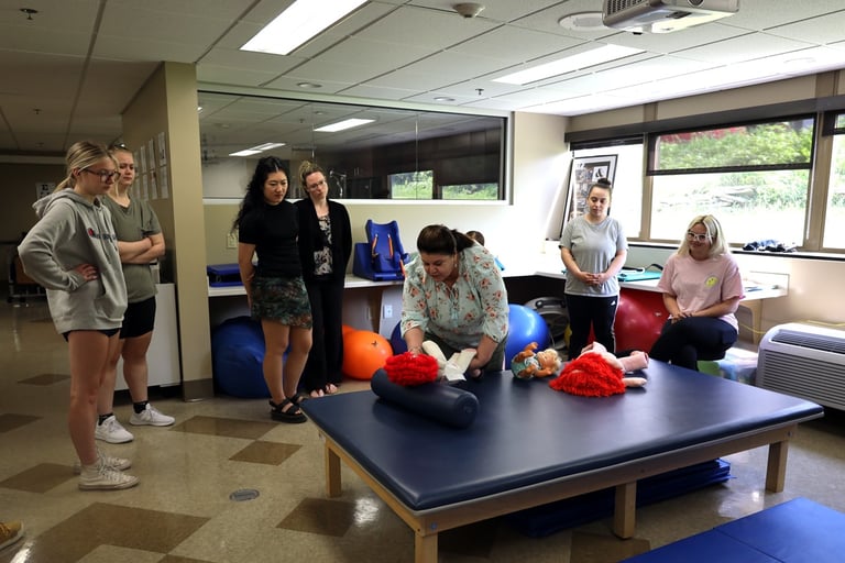 Group of people watching a demonstration on a therapy table with medical equipment and soft toys in a room with large windows.