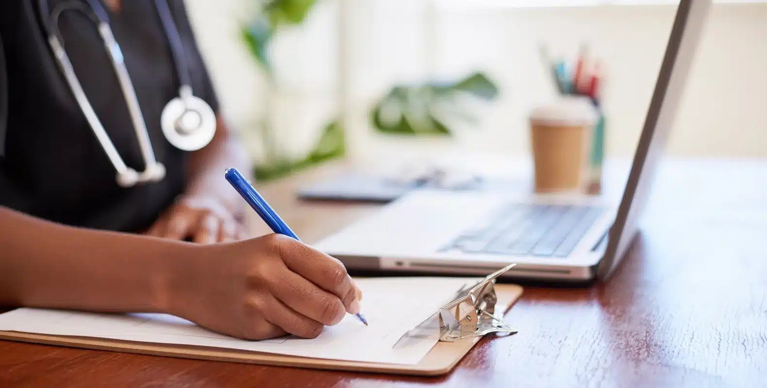 A medical professional writes on a clipboard, with a stethoscope around their neck and a laptop on the desk.