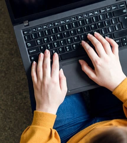 A person wearing a mustard yellow sweater types on a black laptop. The photo is taken from above, focusing on their hands. The laptop has a black keyboard, and the person is seated on a dark surface.