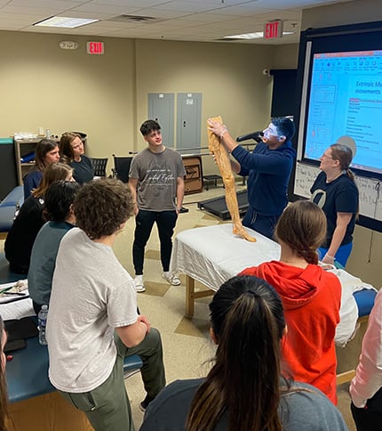 A group of students gathers around a table as an instructor in the PTA Program holds up a preserved human leg for anatomical study during Preview Day in a classroom with a presentation projected on a screen.