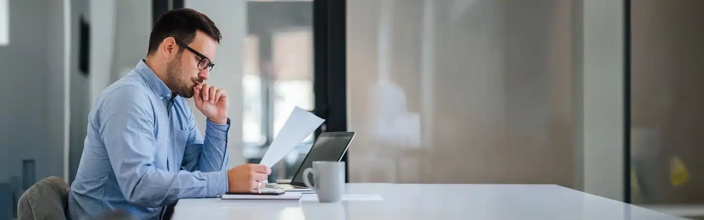 A man in a blue shirt, seated at a desk, deeply focused on a document, with a laptop and coffee mug beside him.