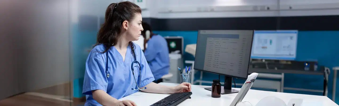 A healthcare worker in scrubs looks at a computer screen in a medical office, with another worker in the background.