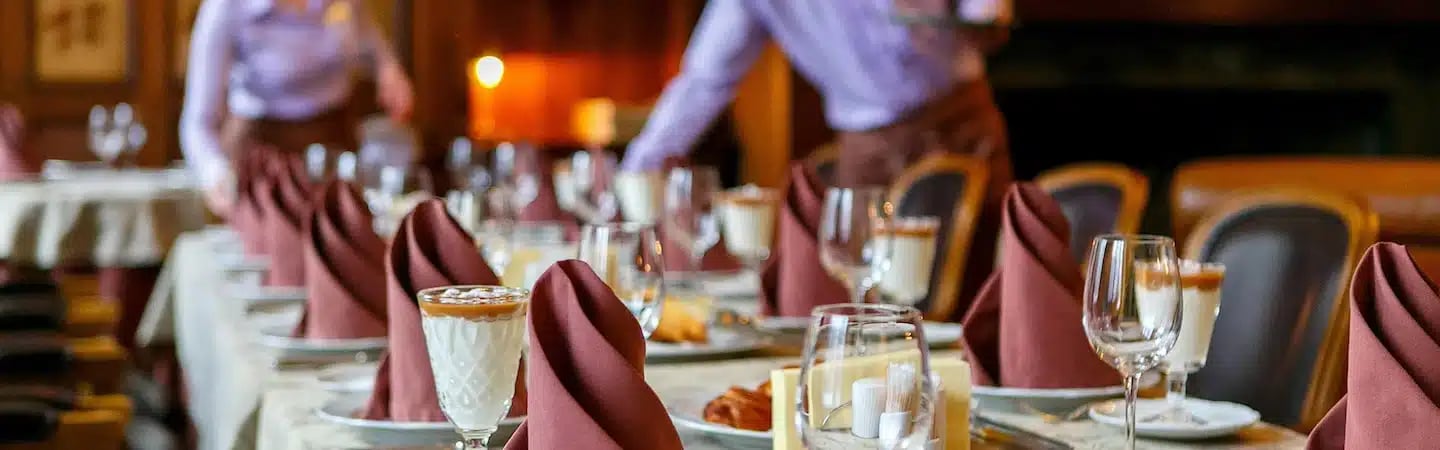 A restaurant dining area with tables set with maroon napkins and glassware, and servers in the background.