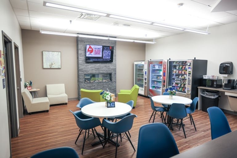A break room in Henrietta, NY, with round tables, blue chairs, vending machines, a microwave, a coffee maker, armchairs, and a TV mounted above a fireplace. Wooden floor and bright lighting.
