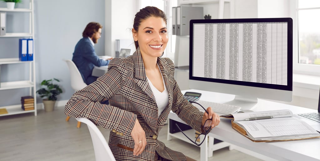 A woman in a plaid blazer sits at a desk, smiling at the camera, holding glasses. Behind her is a computer monitor displaying a spreadsheet—an inviting scene for those looking to enter the field of accounting careers. Another person works in the background.