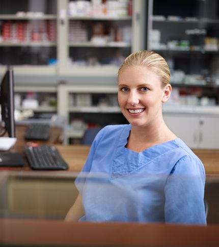 A woman wearing blue medical scrubs sits at a desk in a pharmacy or medical office, smiling at the camera. As a healthcare administrative assistant, she works among shelves filled with medications and supplies in the background.