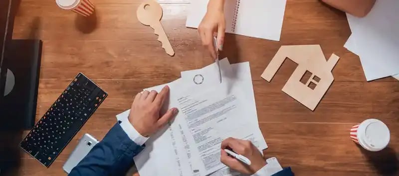 Overhead view of people working at a table with documents, a house model, a key cutout, a keyboard, and coffee cups.
