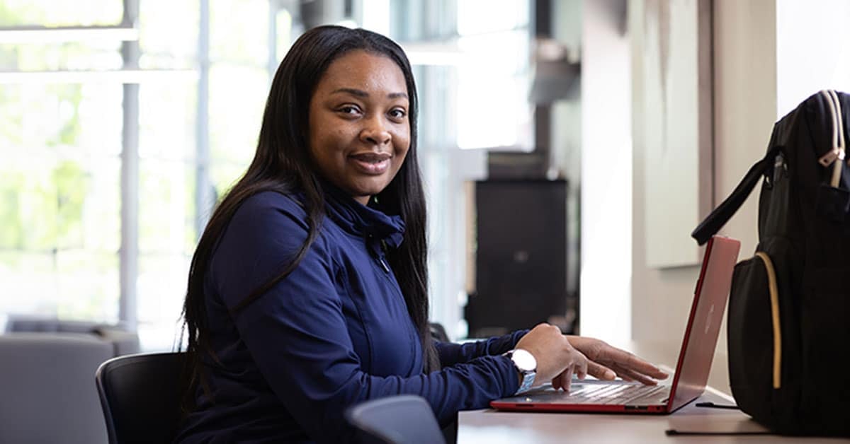 Woman sitting at a desk, smiling at the camera while using a red laptop, with a backpack nearby.