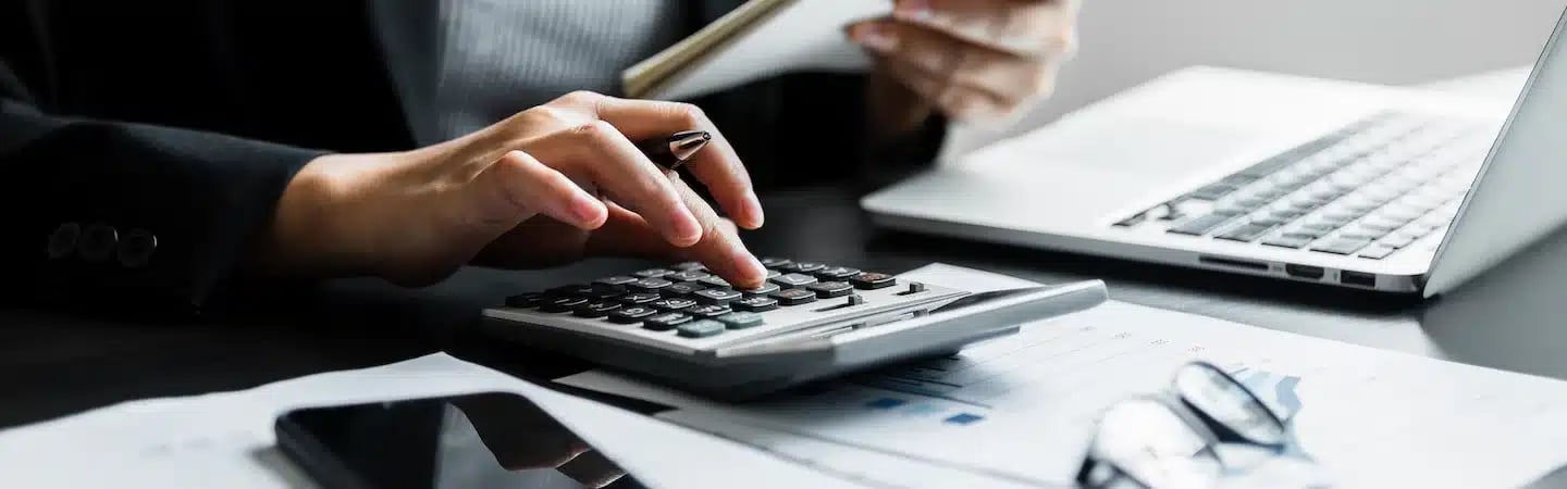 A person using a calculator with financial documents, a notebook, and a laptop on a desk.