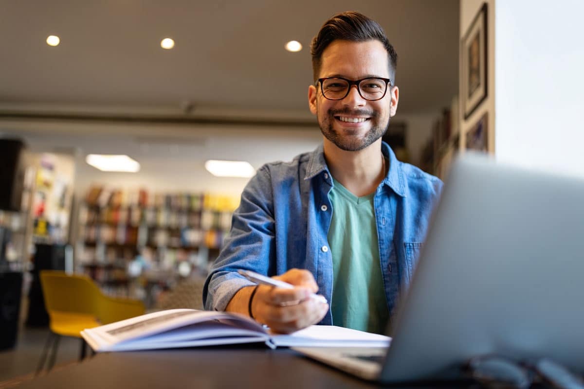 Young man with glasses smiling, holding a pen, and sitting in front of a laptop with an open book in a cozy library.