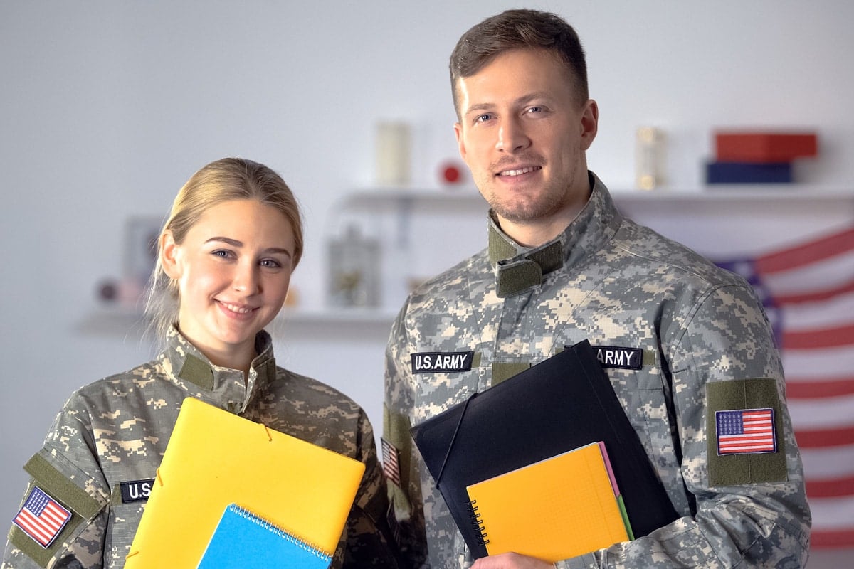 Two U.S. Army soldiers in uniform holding notebooks, smiling in front of an American flag backdrop indoors.