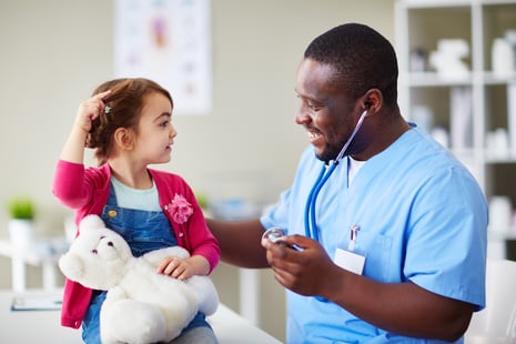 A young girl holding a stuffed bear sits on an exam table while a smiling healthcare professional in blue scrubs and a stethoscope—who recently completed HR Degrees—interacts with her in a medical office.