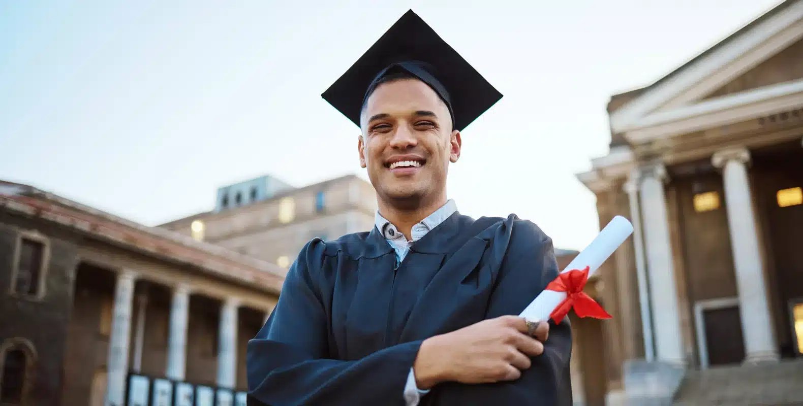 A graduate in cap and gown smiles while holding a diploma, with historic buildings in the background.